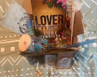 May include: A box filled with various items, including a book titled "Scandalous Lover" by Lore Townsend, a glass with a blue and green tropical design, a straw, a small bag of brown powder, a small bottle with a cork stopper, and a small card with a starfish design.