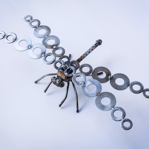 Puede incluir: Una escultura de libélula de metal hecha con arandelas. La libélula tiene un cuerpo y alas plateadas, y una cabeza y cola marrones.
