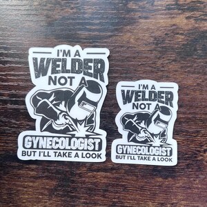 May include: Two white stickers with black text and illustrations. The stickers read "I'M A WELDER NOT A GYNECOLOGIST BUT I'LL TAKE A LOOK." One sticker is larger than the other. The illustrations depict a welder.