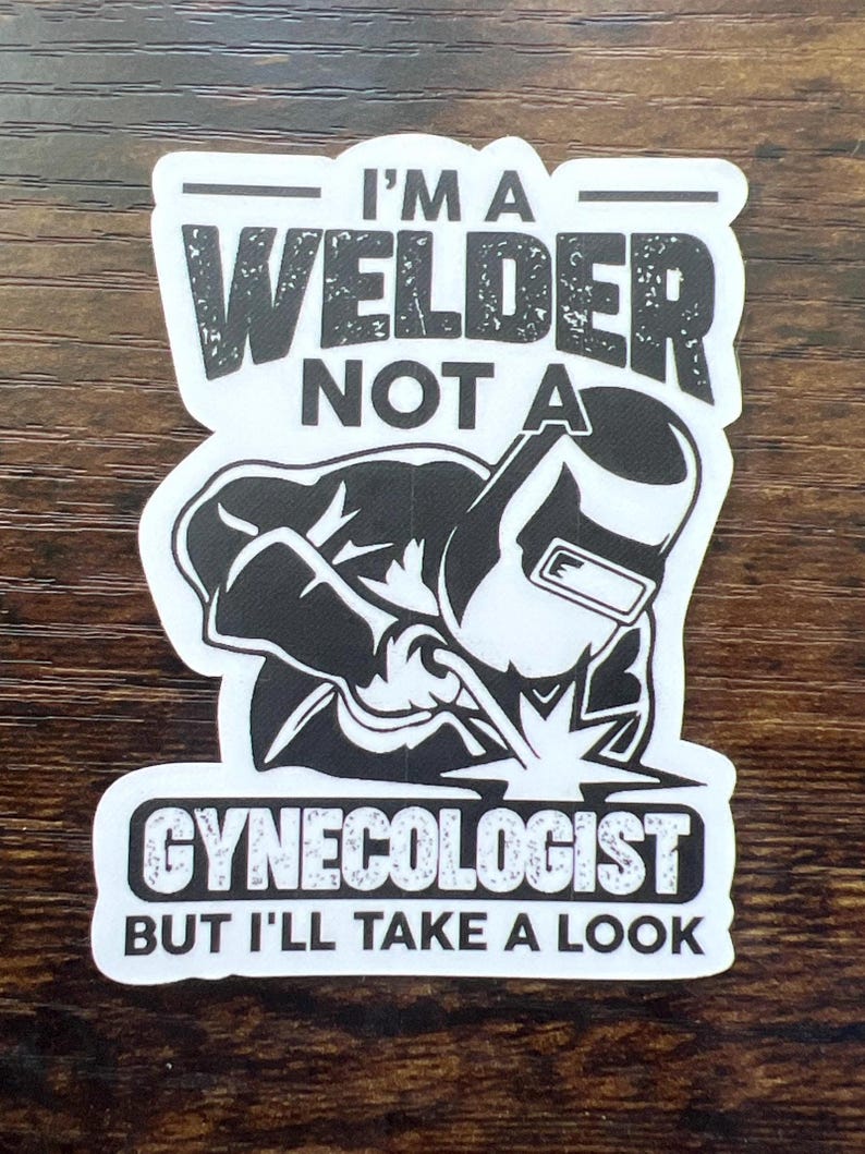 May include: A white sticker with black text and a graphic of a welder. The text reads "I'M A WELDER NOT A GYNECOLOGIST BUT I'LL TAKE A LOOK". The sticker is on a brown wooden surface.