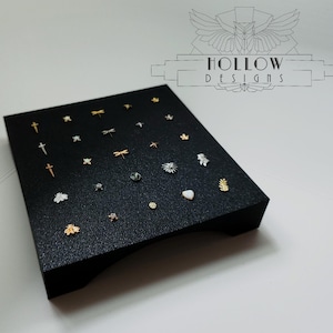 May include: A black display case containing a selection of small gold and silver jewellery pieces, including crosses, stars, dragonflies, and leaves. The case is labelled "HOLLOW DESIGNS".