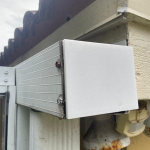 May include: A white, rectangular awning attached to a building's exterior. The awning has a ribbed, metallic underside and a solid white end piece. The structure is mounted near a window and a gutter system, with a weathered appearance.