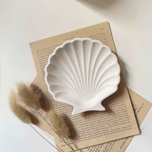 May include: A white ceramic seashell-shaped dish with a textured surface. The dish is sitting on a brown paper background with text.