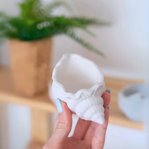Pode incluir: Vaso de cerâmica branco em forma de concha do mar com um interior liso e arredondado. O vaso é segurado em uma mão com esmalte de unha azul claro.