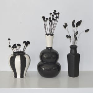 Op de afbeelding: Drie zwart-witte keramische vazen met gedroogde bloemen. De vaas links heeft zwart-witte strepen. De middelste vaas is een zwart-witte kalebasvorm. De vaas rechts is een zwarte rechthoekige vaas.
