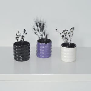 May include: Three small ceramic planters with black rims, each with a different color glaze: black, purple, and white. The planters are arranged in a row on a white surface and each contains dried black flowers.