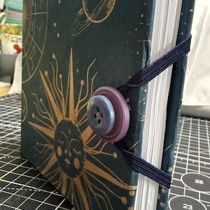 May include: A handmade journal with a celestial design. The cover is dark teal with gold illustrations of planets, suns, and zodiac symbols. It features a navy blue elastic closure and two-tone button. The journal's pages are white.