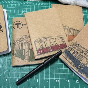 May include: A collection of kraft paper notebooks featuring illustrations of public transportation vehicles. The notebooks depict a bus, subway cars, and the Boston transit logo. A black pen rests on the green cutting mat.