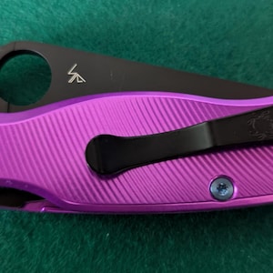 May include: A purple folding knife with a black blade. The handle is a vibrant purple with a textured pattern. The blade has a circular opening and a small logo. The knife has a black pocket clip and blue screws.