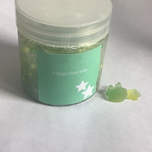Puede incluir: Un tarro transparente con tapa verde y una etiqueta verde que dice "froggy clear jelly". El tarro está lleno de una sustancia gelatinosa verde. Hay dos ranas gomosas verdes al lado del tarro.