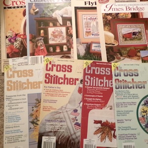 May include: A collection of vintage cross-stitch magazines, featuring colorful covers with various designs. Titles include "The Cross Stitcher" and "Imes Bridge". The magazines are arranged on a neutral surface.