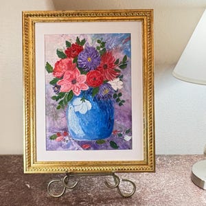 Może przedstawiać: Oprawiony obraz przedstawiający kompozycję kwiatową w niebieskim wazonie. Kwiaty są w odcieniach czerwieni, różu, fioletu i bieli. Obraz jest w złotej ramie i stoi na ozdobnym stojaku.