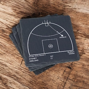 May include: Four square slate coasters stacked on top of each other. Each coaster has a white diagram of a sports field, with the text "Franklin" and "Buddy's 1000th". The coasters are dark grey and sit on a wooden surface.