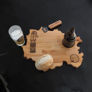 Könnte beinhalten: Holzservierbrett in Form einer Karte, mit einem Bierglas, einem Brötchen und einer Flasche. Das Brett hat ein geschnitztes Design einer Grubenlampe und das Wort "Bochum". Ein Öffner ist ebenfalls enthalten.
