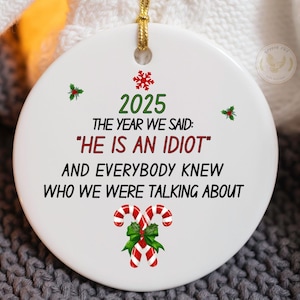 May include: A white ceramic Christmas ornament with gold string. The ornament features the year "2025" in green, the text "THE YEAR WE SAID: 'HE IS AN IDIOT' AND EVERYBODY KNEW WHO WE WERE TALKING ABOUT" in black, and candy cane graphics.