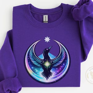 May include: A purple sweatshirt featuring a celestial raven design. The raven is depicted with outstretched wings, set against a backdrop of a crescent moon and a starry galaxy. A white star is above the raven. The sweatshirt is made of a soft material.