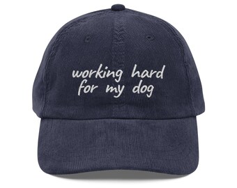 May include: Navy blue corduroy baseball cap with the embroidered text "working hard for my dog" in white. The cap has a curved brim and a top button. A casual accessory for dog lovers.