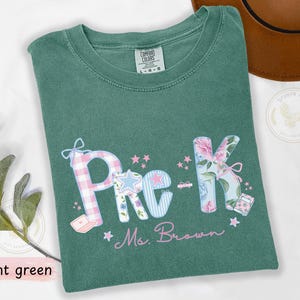 May include: Light green t-shirt featuring the text "Pre-K Ms. Brown". The letters are decorated with plaid, floral, and star patterns. A brown hat is visible in the background.