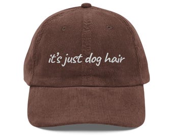 May include: A brown corduroy baseball cap with the embroidered text "it's just dog hair" in white. The cap has a curved brim and a small button on top. The hat is a solid color and has a classic, casual style.