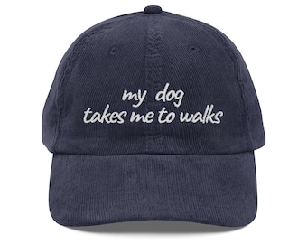 May include: Navy blue corduroy baseball cap with the embroidered text "my dog takes me to walks" in white script. The cap has a curved brim and a top button.