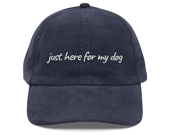 May include: Navy blue corduroy baseball cap with the embroidered text "just here for my dog" in white. The cap has a curved brim and a button on top. A casual accessory for dog lovers.