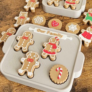 May include: A collection of gingerbread man and cookie-shaped ornaments in a variety of designs. The ornaments are arranged in gray trays with the text "Our Family" on them. Some gingerbread men have red and green accents.