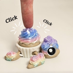 May include: A collection of whimsical, 3D-printed cupcake-shaped objects with colorful frosting in shades of blue, pink, and purple. One cupcake is being pressed by a finger. The word "Click" is written on the image.