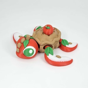 May include: A whimsical 3D-printed apple pie with a miniature apple on top and several apple slices. The pie crust is tan, the apples are red with green leaves, and the slices have white flesh.