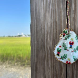 May include: An oyster shell Christmas ornament with a gold-painted edge and a decorative pattern of nutcrackers and greenery. The ornament hangs from a gold and red ribbon against a wooden background.