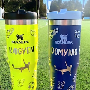 Peut inclure: Deux bouteilles d'eau en acier inoxydable avec poignées. La bouteille de gauche est jaune avec le nom "KAIGYEN" et des graphismes sur le thème du football. La bouteille de droite est bleue avec le nom "DOMYNIQ" et des graphismes sur le thème du football. Les deux bouteilles portent le logo Stanley.