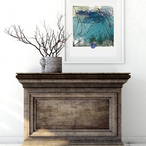 May include: A framed abstract art print with a geometric design in shades of blue, green, and black. The print is hanging on a white wall above a wooden cabinet.