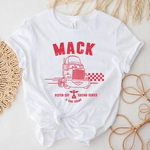 Puede incluir: Camiseta blanca con un gr&aacute;fico rojo de un cami&oacute;n de dibujos animados y la palabra "MACK" encima. El gr&aacute;fico tambi&eacute;n incluye el texto "Piston Cup Racing Series 5 Time Champ". La camiseta est&aacute; anudada a la cintura.