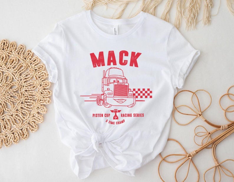 Puede incluir: Camiseta blanca con un gr&aacute;fico rojo de un cami&oacute;n de dibujos animados y la palabra "MACK" encima. El gr&aacute;fico tambi&eacute;n incluye el texto "Piston Cup Racing Series 5 Time Champ". La camiseta est&aacute; anudada a la cintura.