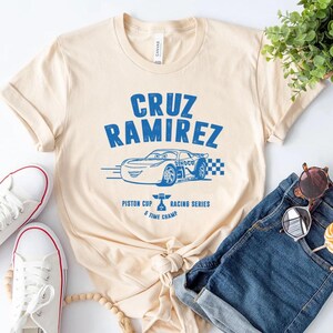 Puede incluir: Camiseta color crema con un gr&aacute;fico azul de un coche de carreras y el texto "CRUZ RAMIREZ". La camiseta tambi&eacute;n incluye el texto "Piston Cup Racing Series" y "5 Time Champ". Zapatillas blancas y pantalones cortos vaqueros tambi&eacute;n en la imagen.