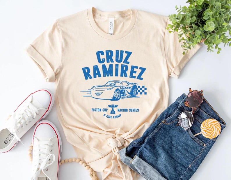 Puede incluir: Camiseta color crema con un gr&aacute;fico azul de un coche de carreras y el texto "CRUZ RAMIREZ". La camiseta tambi&eacute;n incluye el texto "Piston Cup Racing Series" y "5 Time Champ". Zapatillas blancas y pantalones cortos vaqueros tambi&eacute;n en la imagen.