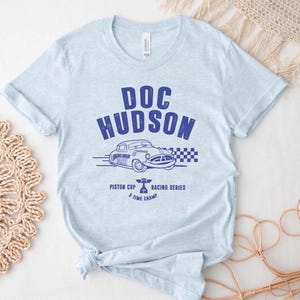 Puede incluir: Camiseta azul claro con un gr&aacute;fico azul de un coche cl&aacute;sico y el texto "DOC HUDSON". El dise&ntilde;o incluye "Piston Cup Racing Series" y "5 Time Champ". La camiseta es de un material suave.