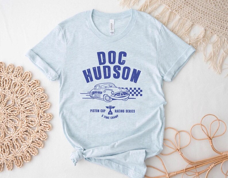 Puede incluir: Camiseta azul claro con un gr&aacute;fico azul de un coche cl&aacute;sico y el texto "DOC HUDSON". El dise&ntilde;o incluye "Piston Cup Racing Series" y "5 Time Champ". La camiseta es de un material suave.