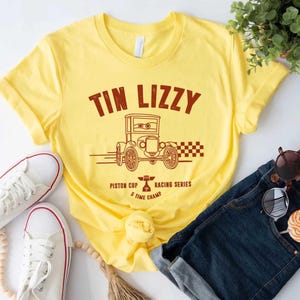 Puede incluir: Camiseta amarilla con un gr&aacute;fico marr&oacute;n de un coche antiguo y el texto "TIN LIZZY". La camiseta tambi&eacute;n presenta el texto "Piston Cup Racing Series 5 Time Champ". Zapatillas blancas y vaqueros azules tambi&eacute;n est&aacute;n en la imagen.