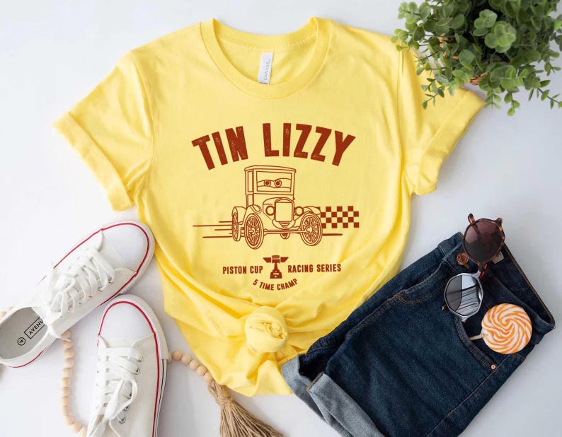 Puede incluir: Camiseta amarilla con un gr&aacute;fico marr&oacute;n de un coche antiguo y el texto "TIN LIZZY". La camiseta tambi&eacute;n presenta el texto "Piston Cup Racing Series 5 Time Champ". Zapatillas blancas y vaqueros azules tambi&eacute;n est&aacute;n en la imagen.