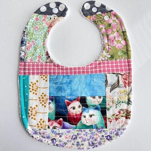 May include: A patchwork baby bib with a variety of fabric patterns. The bib features a central panel with a cat print, surrounded by floral, polka dot, and plaid fabrics. The bib is secured with snaps.
