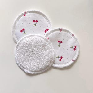 May include: Three reusable facial rounds. Two rounds feature a white fabric with a cherry print in red and green. The third round is white terry cloth. The rounds are circular and designed for skincare.