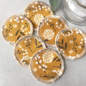 May include: Reusable, round facial cleansing pads. The pads feature a mustard yellow floral pattern with white and black accents. Each pad has a stitched, light brown border. A glass jar is visible in the background.