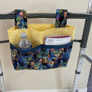 May include: A quilted walker bag featuring a colorful cat print with blue, green, and yellow tones. The bag has two top straps, a yellow interior, and a dark blue front pocket containing a clear plastic water bottle and a booklet titled The Word.