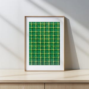 以下が含まれることがあります： 緑と黄色の格子模様の額入りアートワーク。格子は、黄色の線が入った濃い緑色の正方形で構成されています。アートワークは金のフレームに入っており、木製の表面に置かれています。