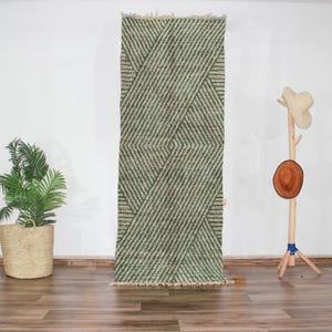 Puede incluir: Una alfombra rectangular verde y beige con un patrón geométrico de líneas diagonales. La alfombra tiene una franja en la parte inferior y se muestra verticalmente. Una planta en maceta y un perchero están en el fondo.