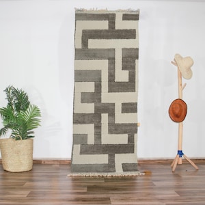 Peut inclure: Un tapis géométrique blanc et gris à motifs avec une texture tissée. Le tapis est suspendu à un mur blanc.