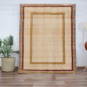 Peut inclure: Un tapis beige avec une bordure multicolore. Le tapis a une grande bordure intérieure rectangulaire avec une plus petite bordure intérieure rectangulaire. Le tapis est sur un mur blanc.