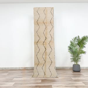 Op de afbeelding: Een wit en crèmekleurig loper met een geometrisch patroon in zwart. Het tapijt is gemaakt van wol en heeft een zachte, pluche textuur.