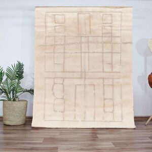 Op de afbeelding: Een crèmekleurig tapijt met een geometrisch patroon van vierkanten en rechthoeken. Het tapijt is gemaakt van een zacht, pluche materiaal en heeft een subtiele glans.