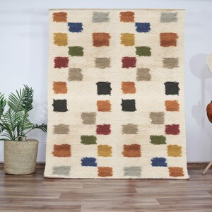 Peut inclure: Un tapis blanc avec un motif géométrique de carrés colorés. Les carrés sont dans des tons de brun, orange, rouge, vert, bleu et noir.